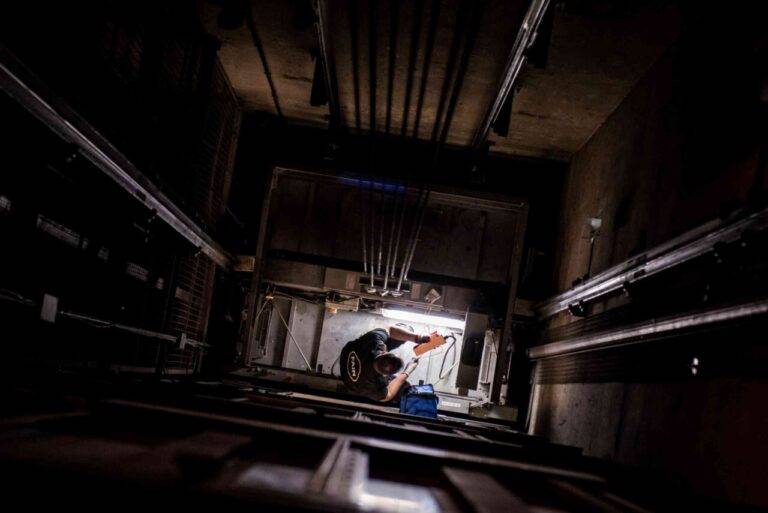 Un technicien de maintenance, équipé de gants, travaille sur des câbles et des équipements dans un espace industriel, avec un sac à outils bleu posé au sol.