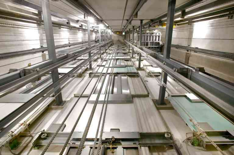 Un technicien debout sur une cabine d'ascenseur dans une cage entourée de câbles et rails, avec une lumière douce illuminant la scène et des panneaux de sécurité en évidence.