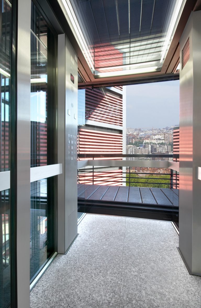 Image montrant une vue d'un ascenseur. Sur le côté gauche, les portes métalliques sont fermées avec des lignes verticales. Sur le côté droit, les portes sont ouvertes, révélant un intérieur métallique avec un sol gris à texture similaire à un tapis.