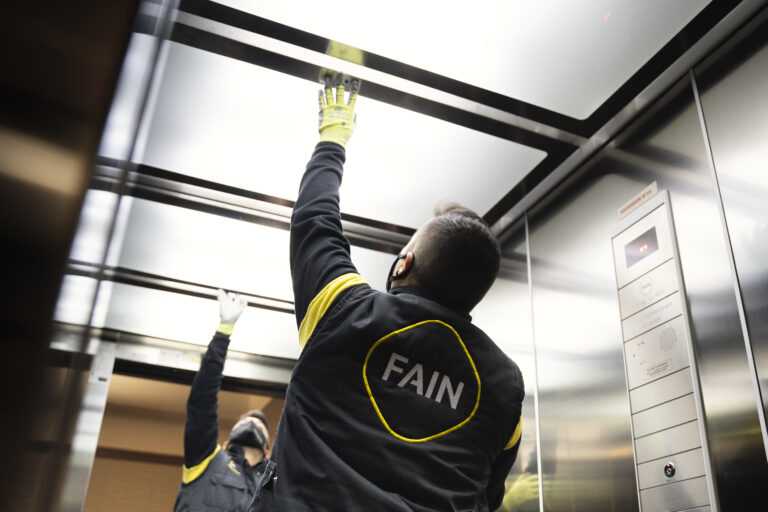 Intérieur d'un ascenseur avec des parois métalliques, un sol texturé et un panneau de commande lumineux comprenant des boutons pour étages, un bouton d'urgence et un haut-parleur.