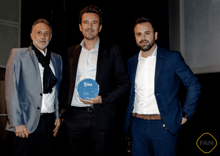 Un design circulaire avec un fond bleu et des lignes géométriques blanches, mettant en avant le prix 'LES TROPHÉES DE L'ASCENSEUR 2022' décerné à FAIN Ascenseurs France pour son partenariat avec les institutions d'inclusion.