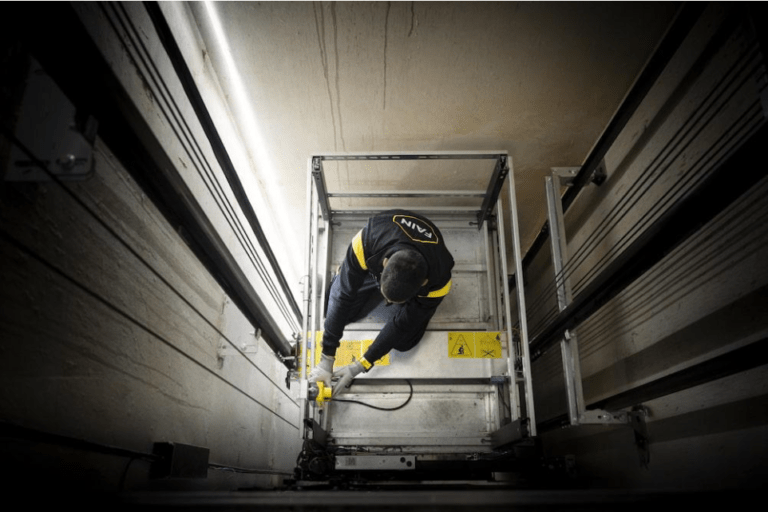 Technicien portant une tenue FAIN, travaillant dans une gaine d’ascenseur avec des outils et des panneaux d’avertissement au sol.