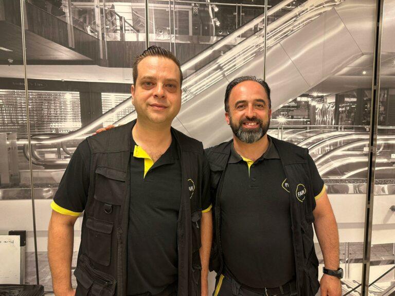 Deux techniciens FAIN souriants assis à une table, dans un cadre intérieur avec plantes et extincteur en arrière-plan.