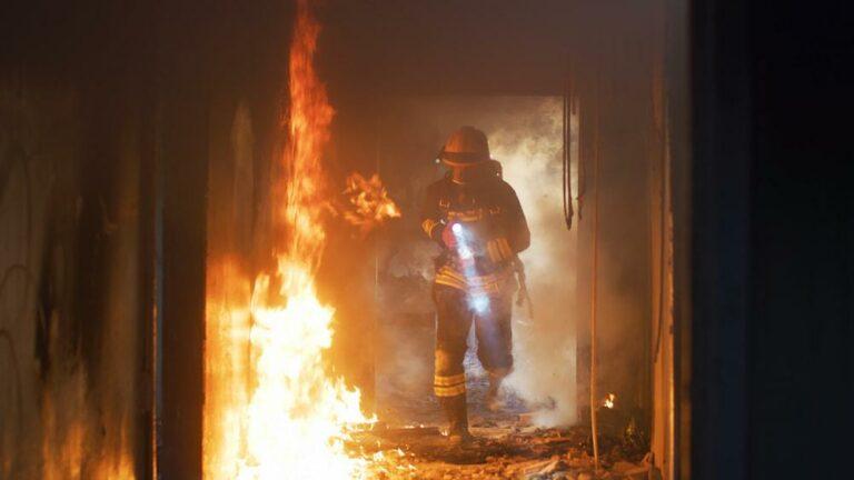 Un pompier dans un bâtiment en feu, équipé de protections et d'une lampe de poche, entouré de flammes et de fumée.